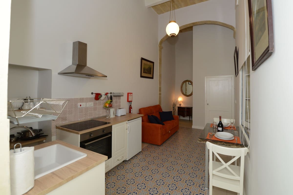 A compact kitchen with a sink, stove, and oven, a dining area with a table and chairs, and a living space with a sofa and a mirror.
