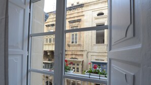 A view through a window with a balcony, featuring a building with classical architecture and a flower box with red flowers.