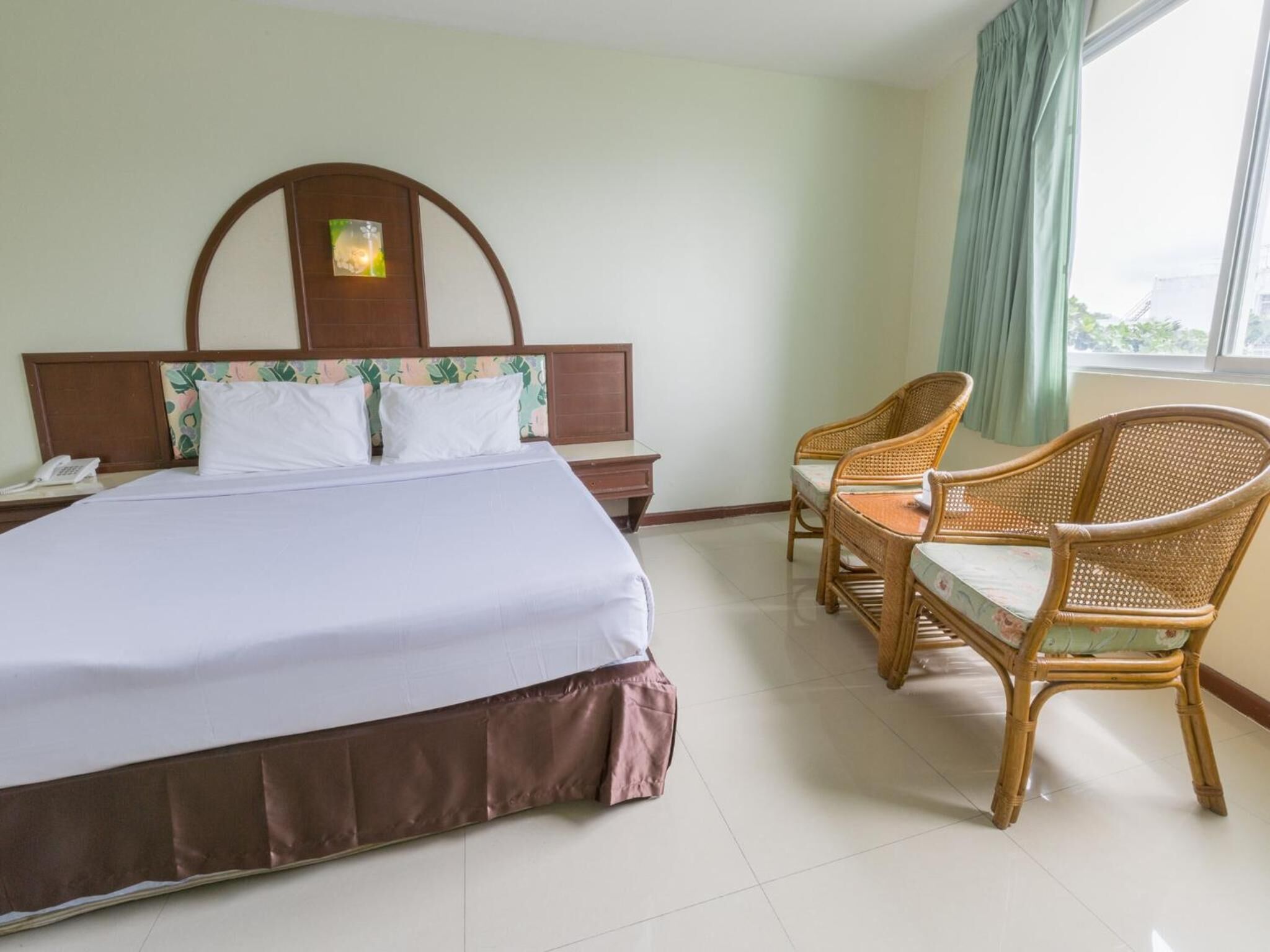 A hotel room with a bed, a nightstand, a telephone, a window with curtains, and two wicker chairs.