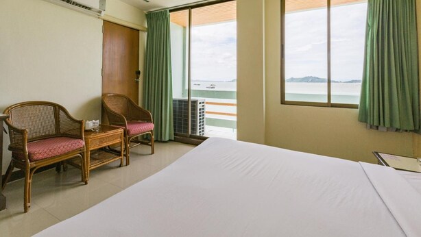 A hotel room with a bed, two wicker chairs, a window with a view of the sea, and an air conditioning unit. - Lake Inn Hotel (Songkhla)