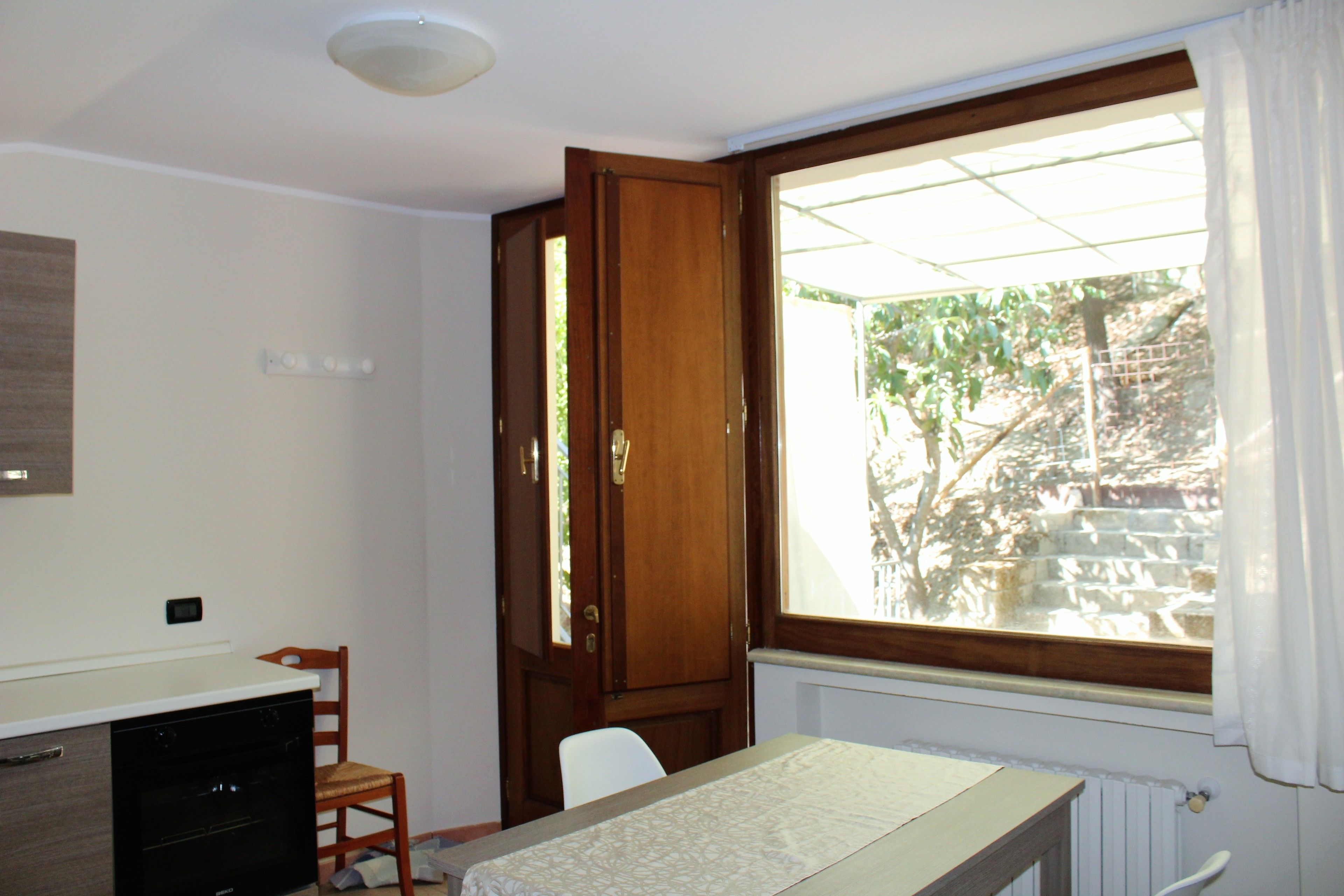 A small room with a wooden door, a window with a view of a garden, a white chair, and a small table.