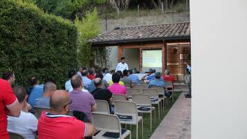 A group of people seated outdoors in a semi-circular arrangement, facing a speaker at a podium.