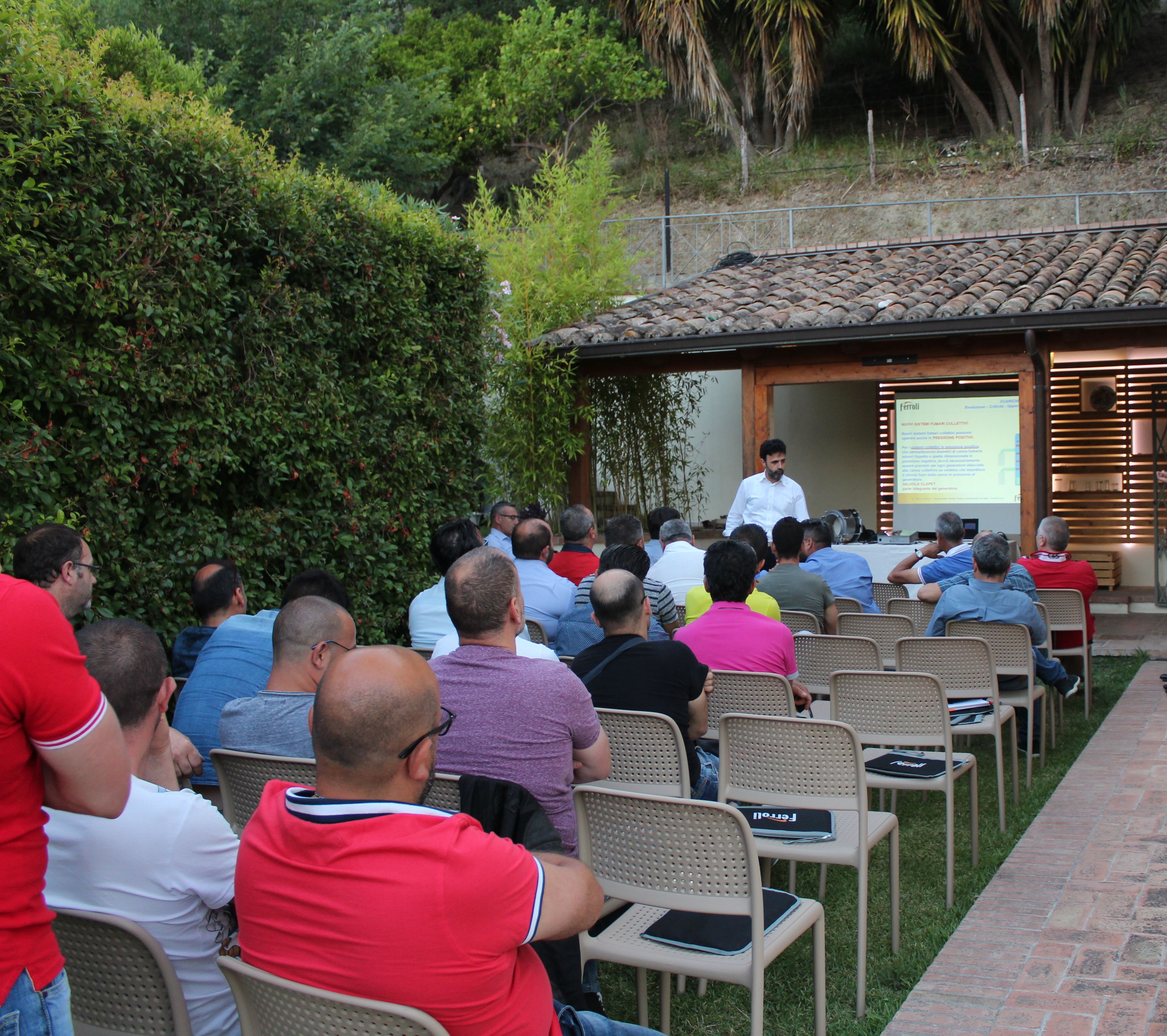 A group of people seated outdoors in a semi-circular arrangement facing a speaker at a podium.