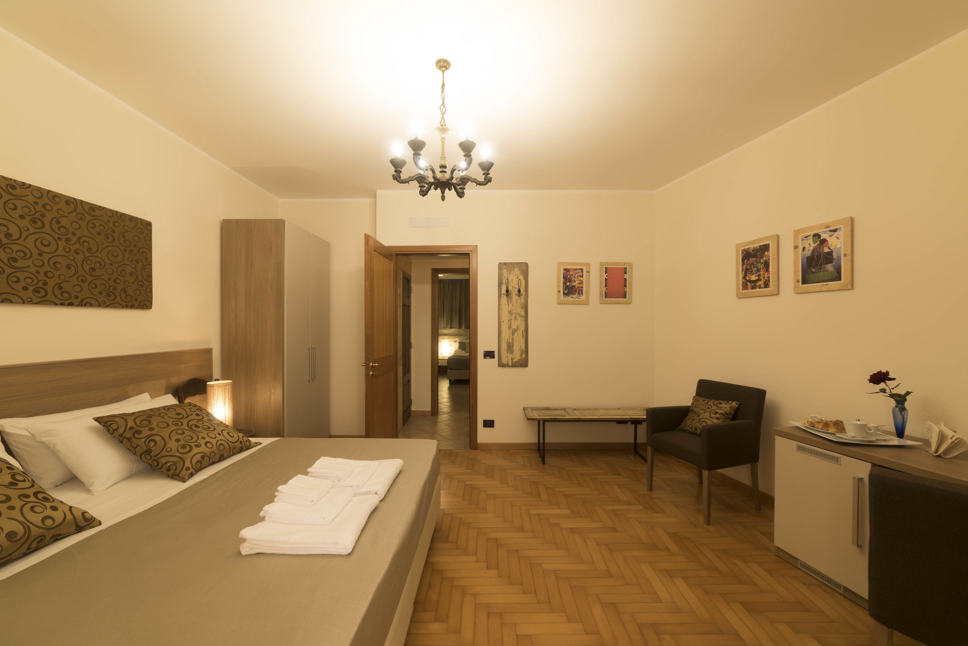 A hotel room with a bed, a desk, a chair, a small kitchenette, and a door leading to another room.