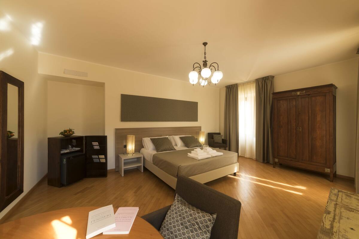 A modern hotel room with a large bed, a desk with papers, a wardrobe, and a chandelier.