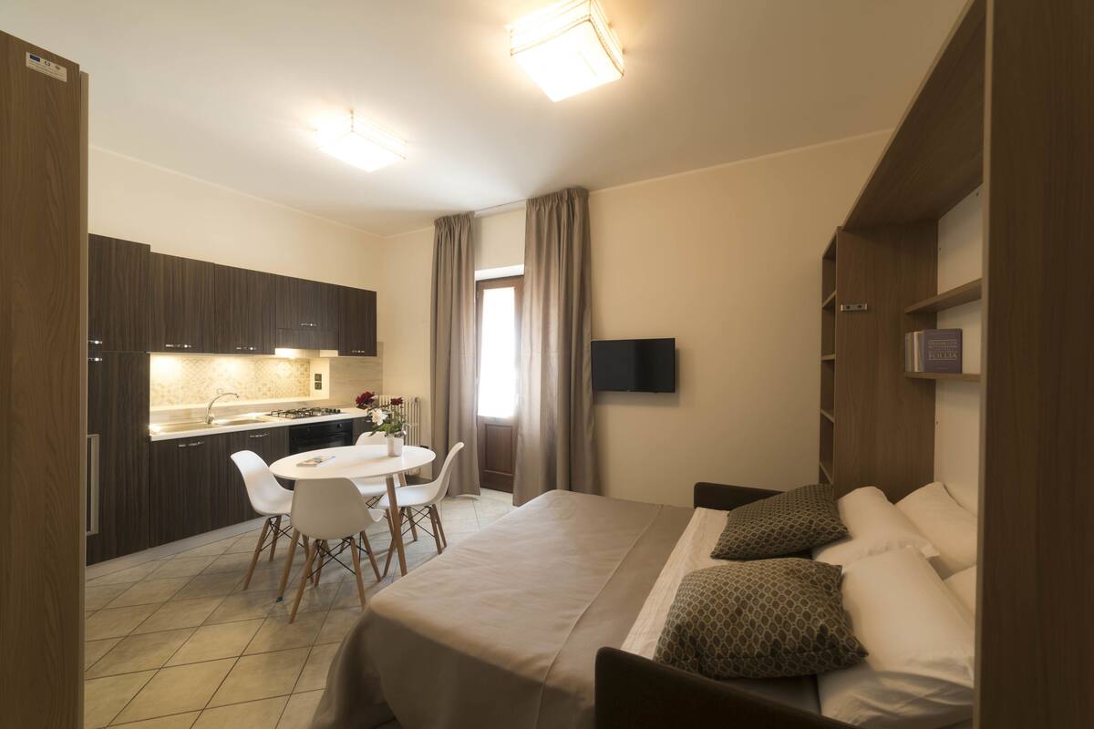 A hotel room with a bed, a desk, a chair, a TV, and a kitchenette.