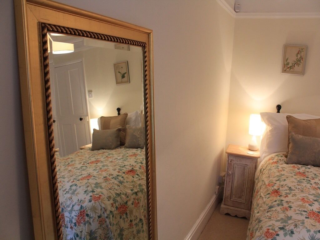 A bedroom with a large mirror, floral bedspread, bedside table with lamp, and a small cabinet.