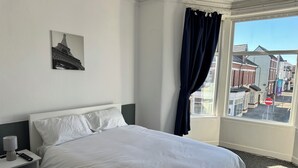 A hotel room with a bed, a nightstand, a lamp, a window with a view of buildings, and a 'WELCOME' sign. - Avari Beach Hotel (Blackpool)
