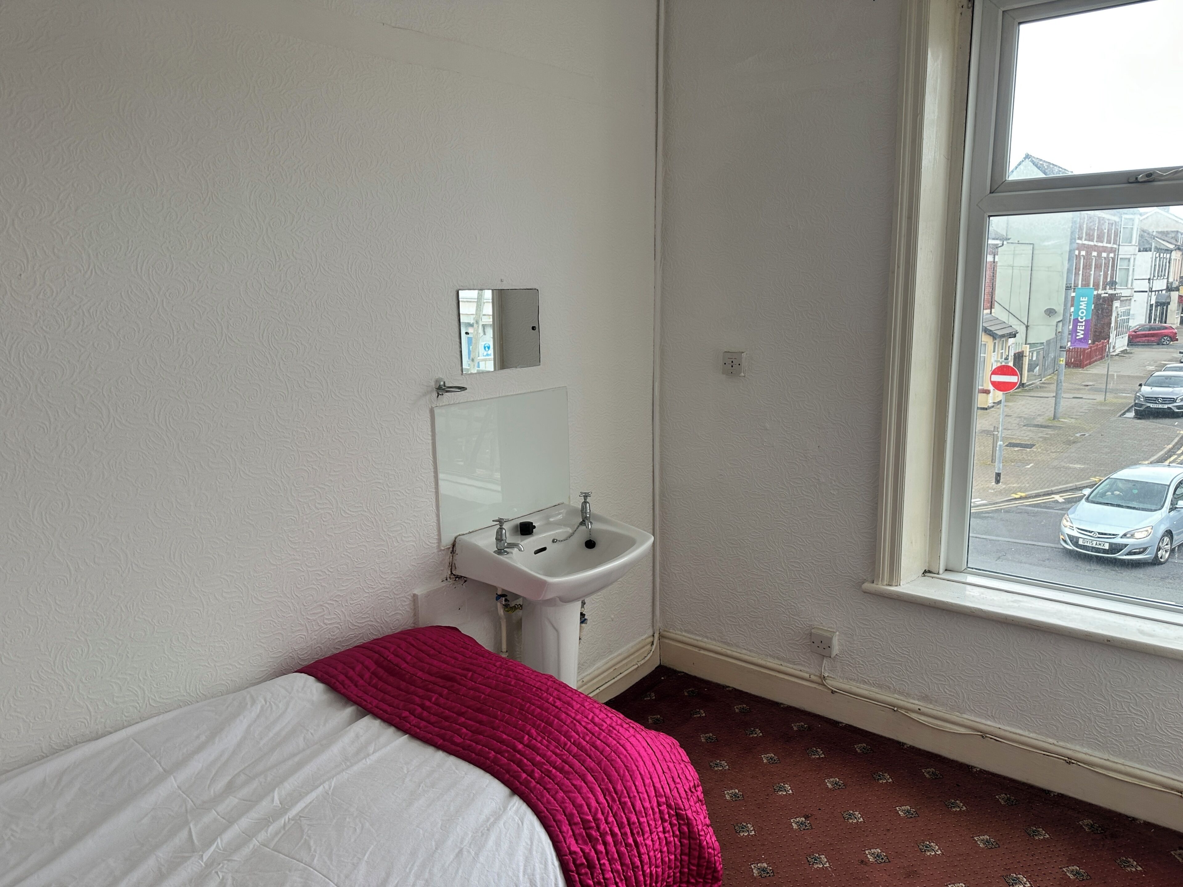 A room with a bed, a window with a view of the street, and a white sink with a mirror.