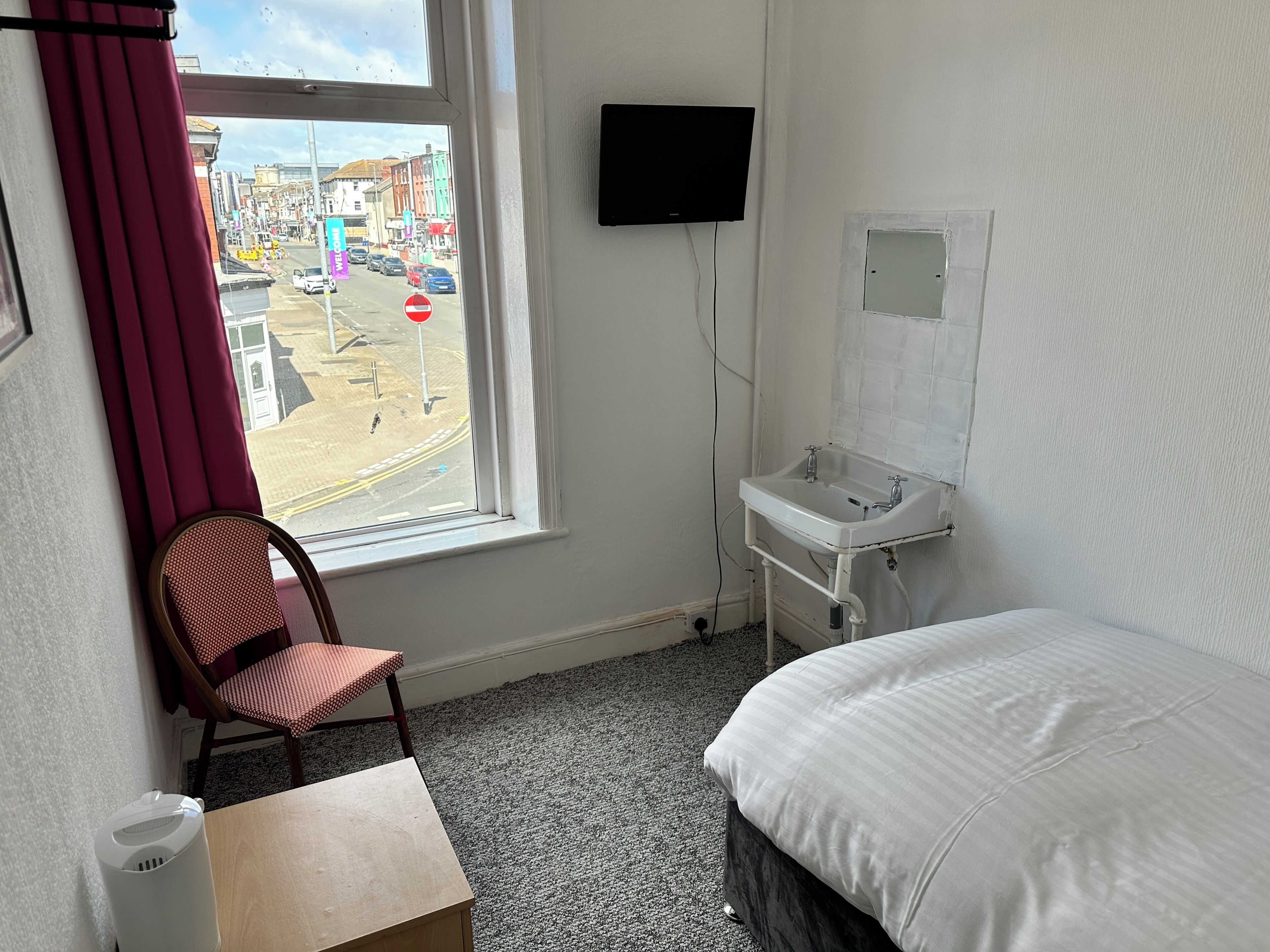 A hotel room with a bed, a desk, a chair, a window with curtains, a TV mounted on the wall, and a sink.
