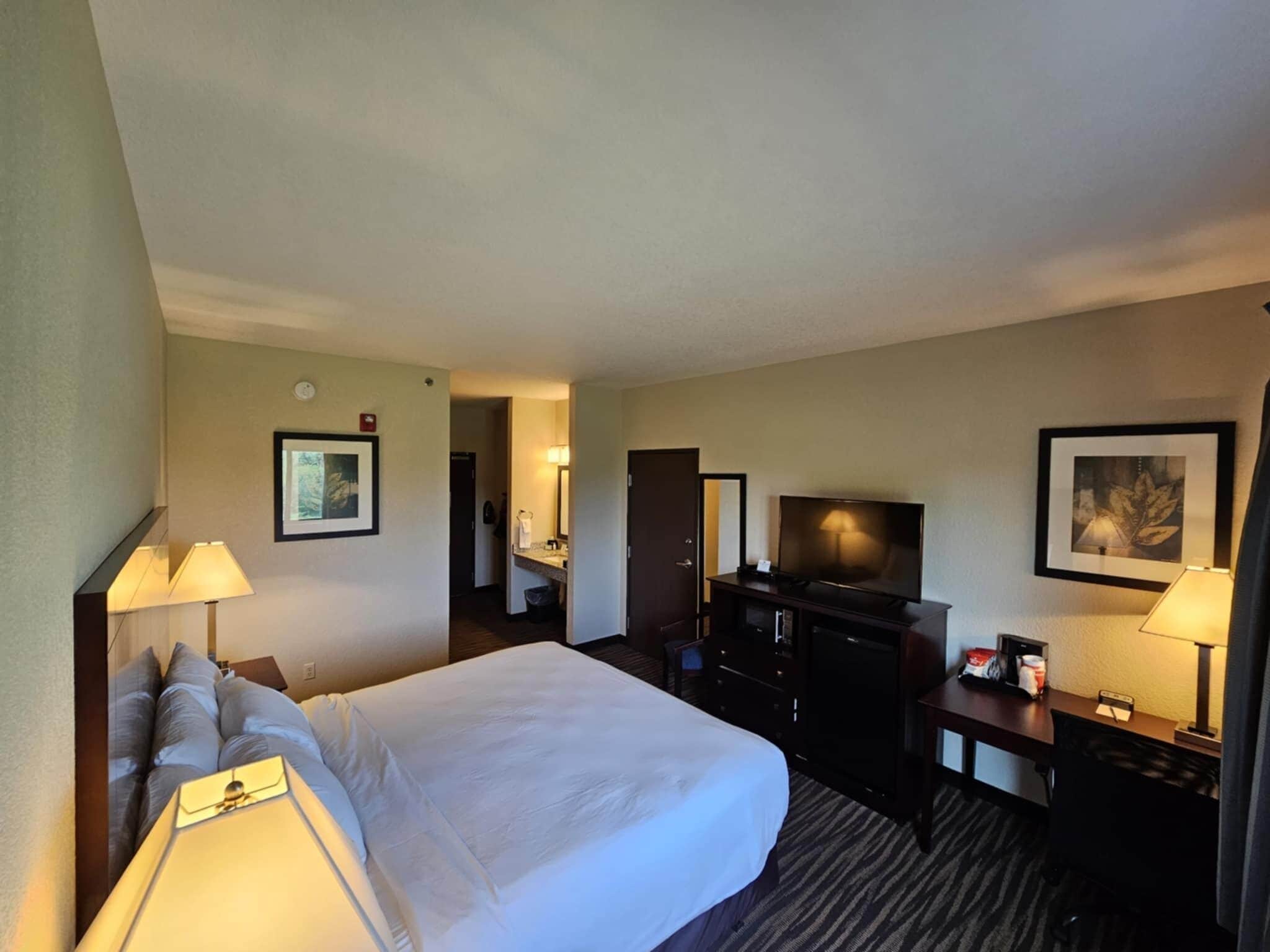 Habitación de hotel con una cama grande, un escritorio con televisor, un aparador y un baño a la vista al fondo.