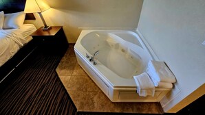 Une chambre d’hôtel dotée d’une grande baignoire, d’un lit avec du linge de lit blanc, d’une table de chevet avec une lampe et d’une tête de lit de couleur sombre.