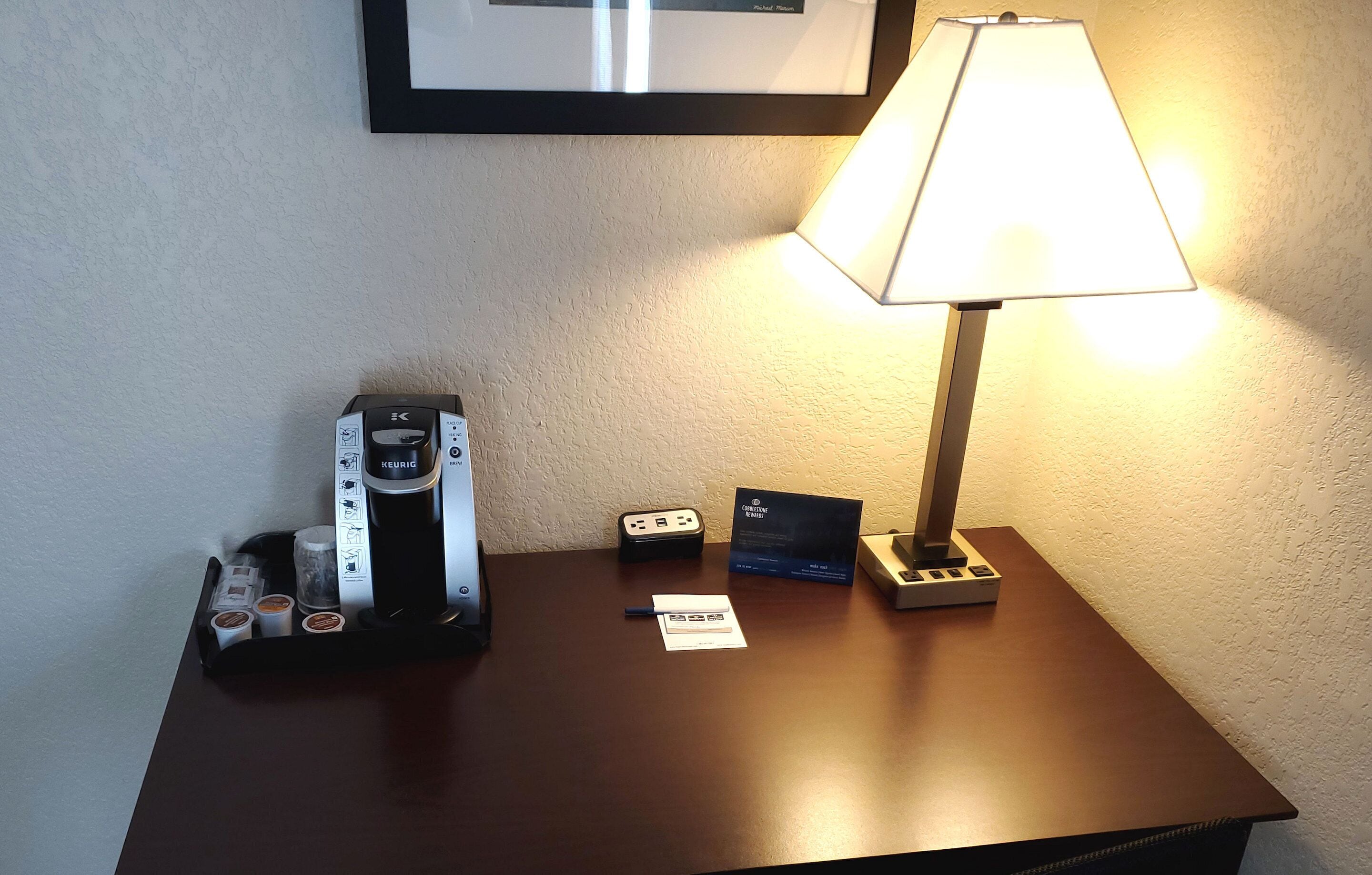 Une chambre d’hôtel équipée d’une machine à café, d’une lampe, d’une multiprise et d’un tableau encadré au mur.