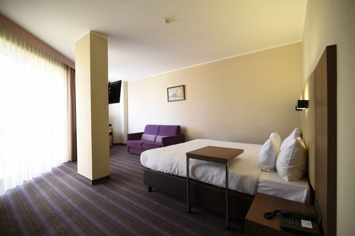 A hotel room with a bed, a small table, a sofa, a TV mounted on the wall, and a window with curtains.