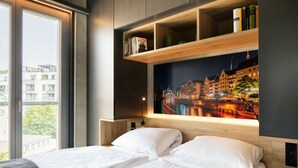 Una habitación de hotel con una cama grande, dos mesitas de noche, una estantería y un cuadro de un paisaje urbano en la pared.
