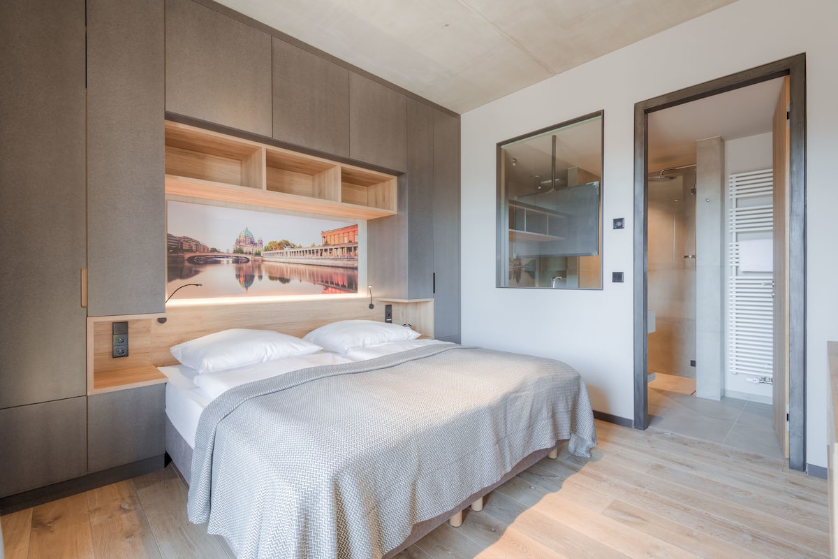 A modern bedroom with a bed, built-in shelving, a large mirror, and a view of a cityscape through the window.