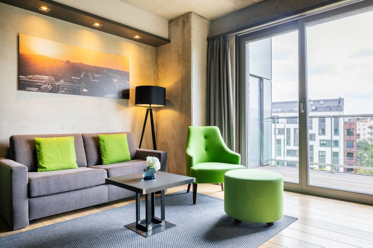 A modern living room with a grey sofa, green cushions, a black floor lamp, a green armchair, and a large window offering a city view.