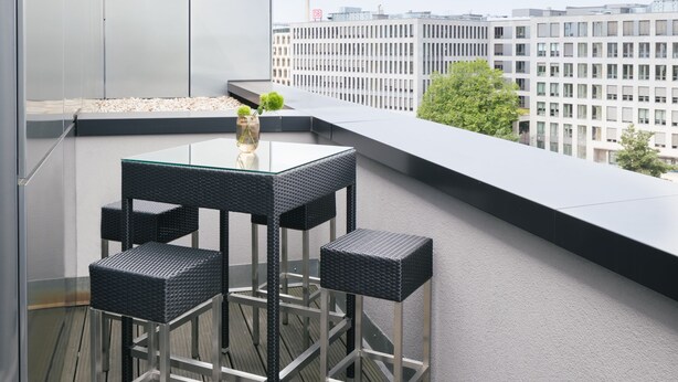 Eine Dachterrasse mit einem Glastisch und vier Korbstühlen, mit Blick auf eine Stadtlandschaft.