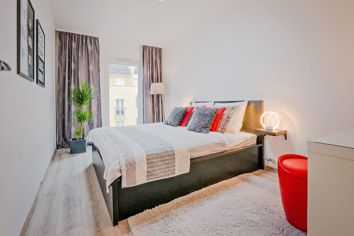 A modern bedroom with a bed, bedside table, lamp, and a red chair.