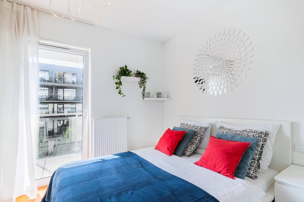 A bedroom with a bed, a window with a view of buildings, a white wall with a decorative item, and a white nightstand.