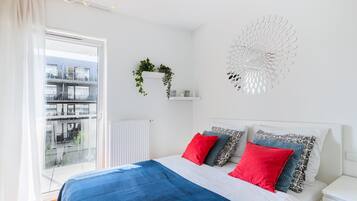 A bedroom with a bed, a window with a view of buildings, a white wall with a decorative item, and a white nightstand.