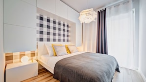A modern bedroom with a checkered headboard, a bedside table with a lamp, and a large window with curtains. - Apartamenty Homely Place City (Poznan)
