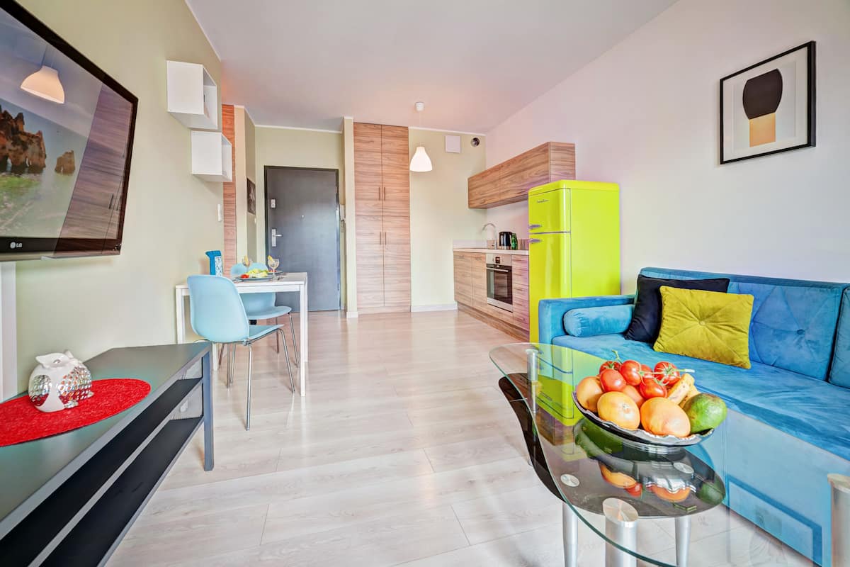 A modern living room with a blue sofa, a glass coffee table with fruit, a dining area with a blue chair, and a kitchen with a green refrigerator.