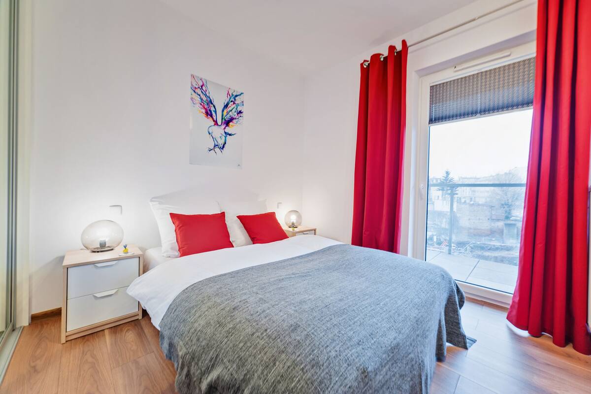 A bedroom with a bed, a nightstand, a window with red curtains, and a city view.