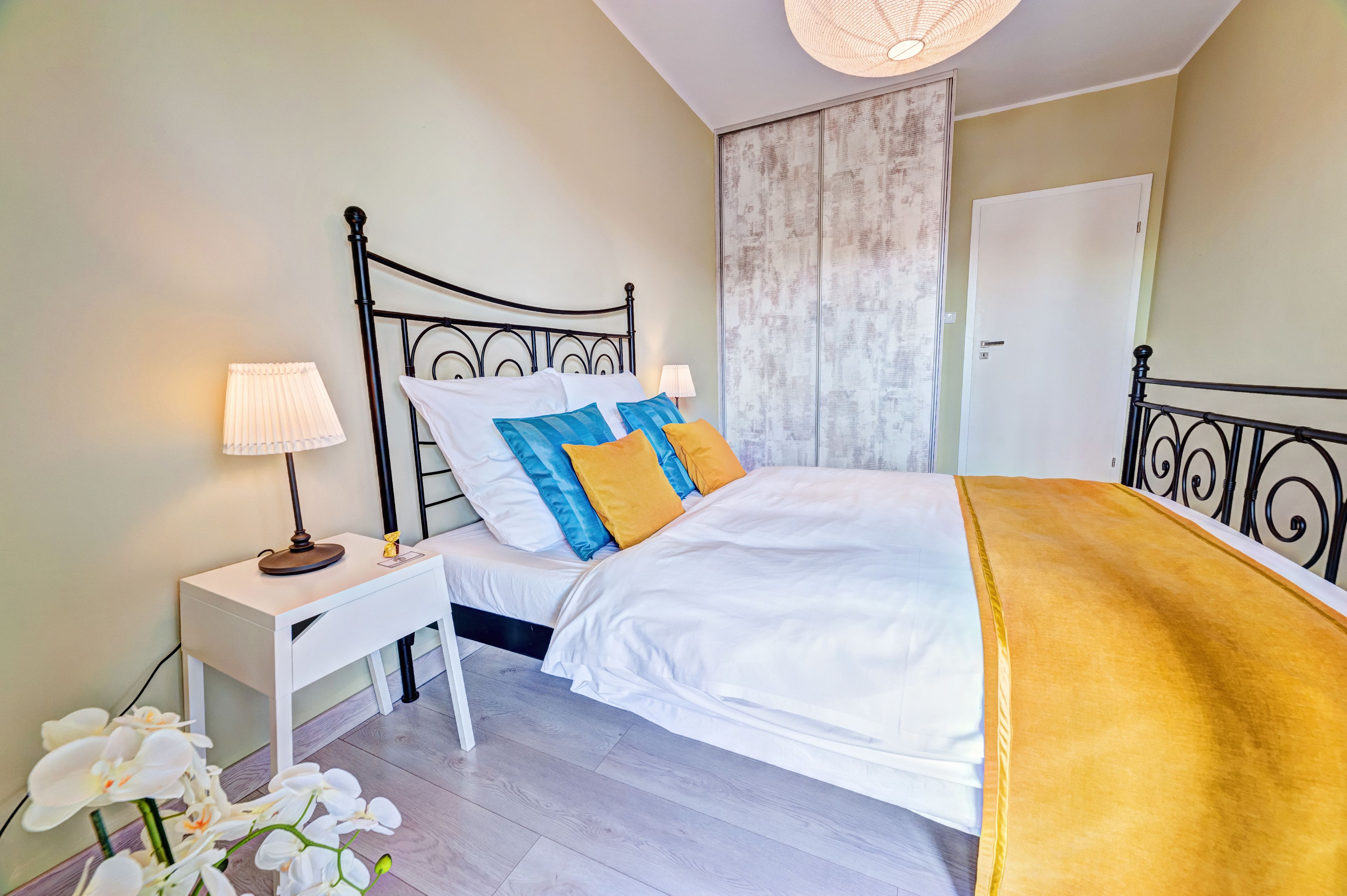 A neatly made bed with white bedding and colorful pillows, a bedside table with a lamp, and a white nightstand.
