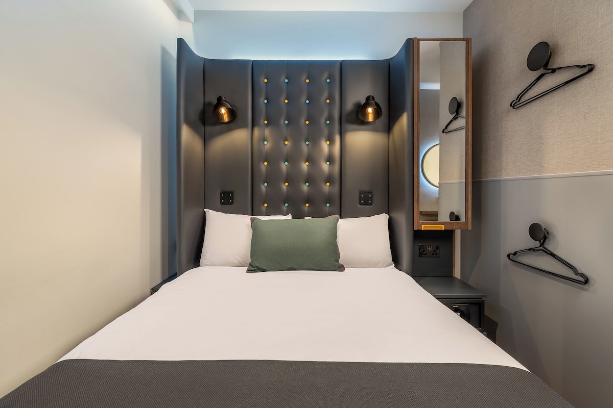Ein modernes Hotelzimmer mit einem großen Bett, einem gesteppten Kopfteil, zwei Wandlampen, einem Spiegel und einem Kleiderhaken.