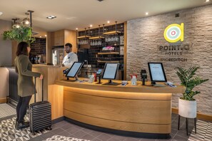 En hotellreception med en bardisk i trä, digitala incheckningskiosker och ett vinställ som är monterat på väggen.