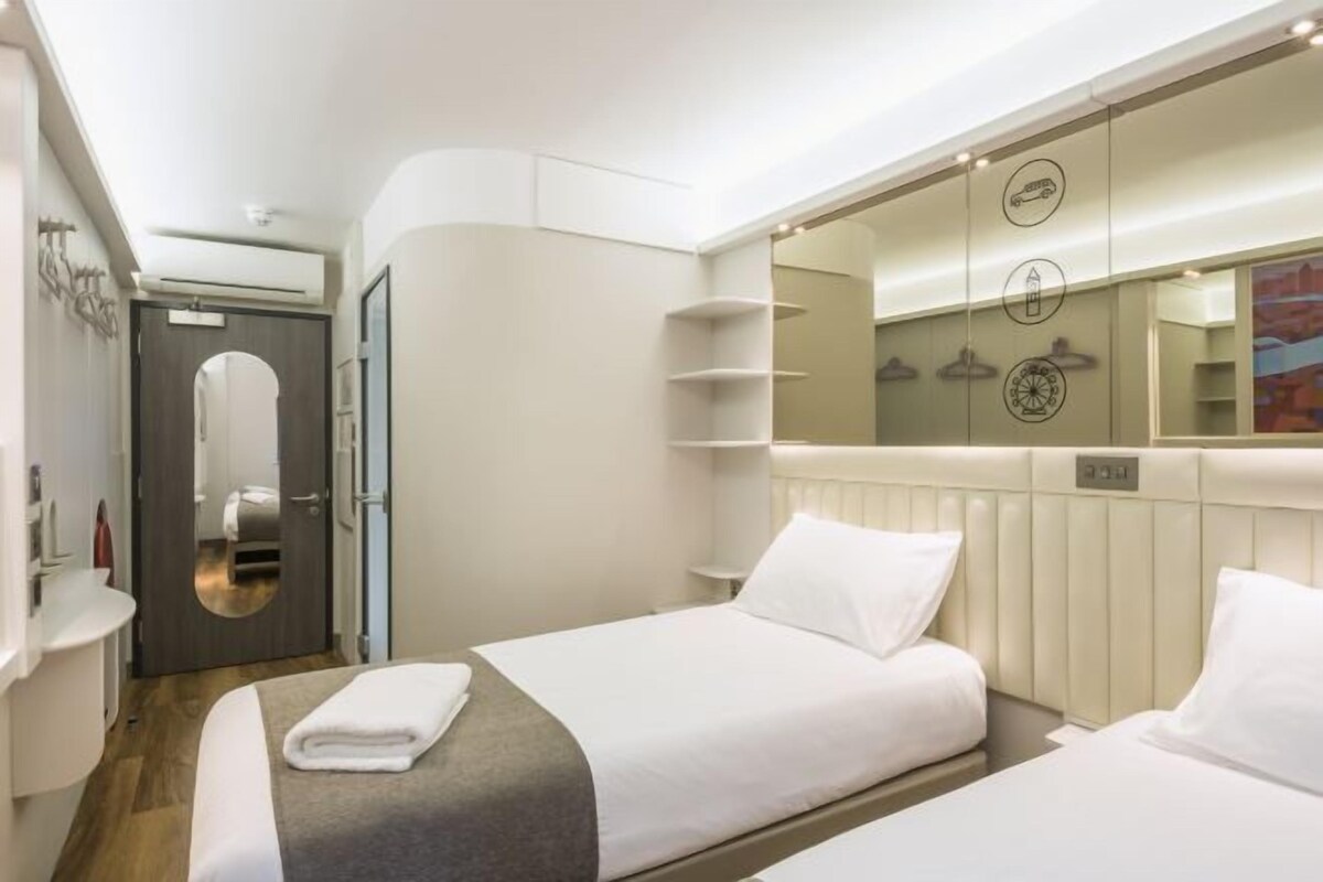 A hotel room with two beds, a large mirror, and a built-in shelf with a clock and a trash bin.