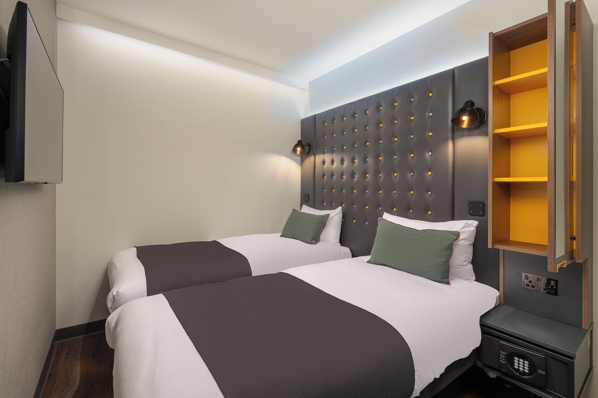 Ein Hotelzimmer mit zwei Betten, einem modernen Kopfteil, Nachttischlampen, einer eingebauten Ablage und einer Bedienkonsole.