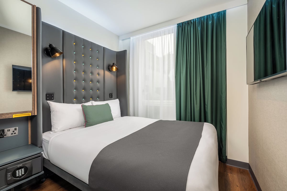 Ein modernes Hotelzimmer mit einem großen Bett, einem mit Beleuchtung versehenen Kopfteil, einem Nachttisch mit Telefon und einem großen Fenster mit Vorhängen.