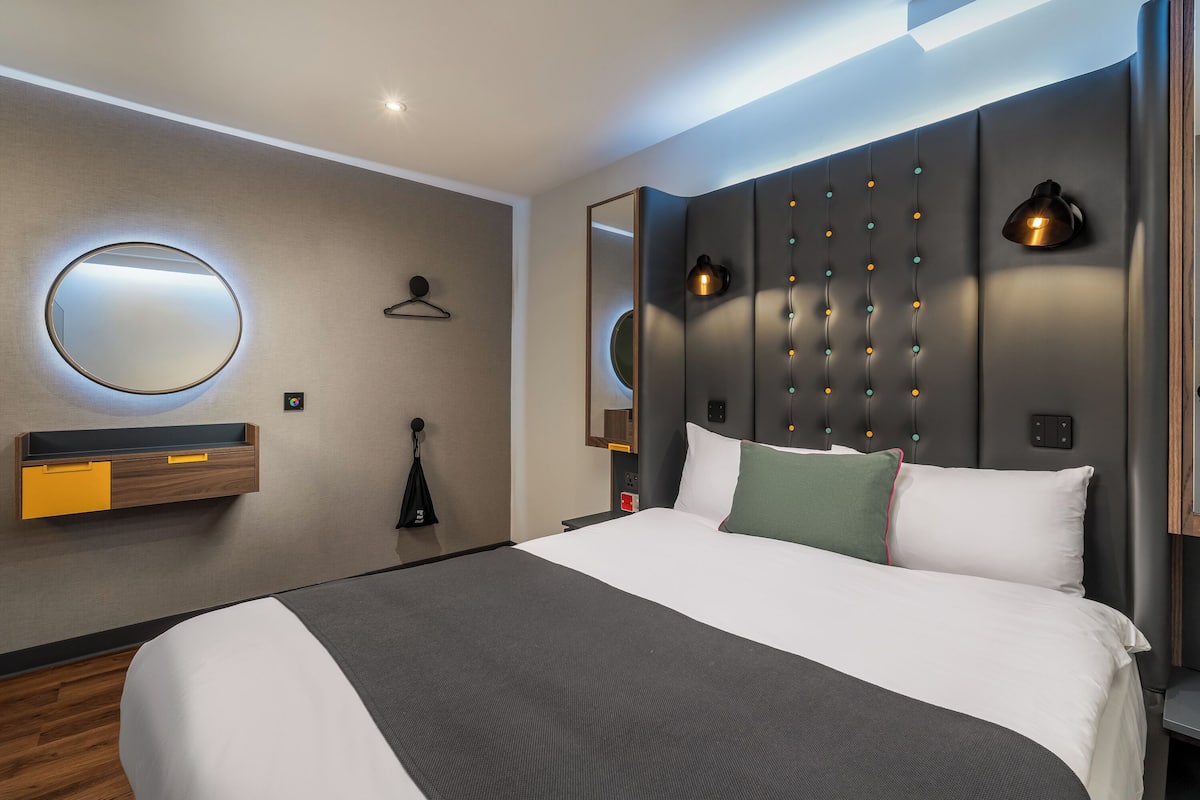 Ein modernes Hotelzimmer mit einem großen Bett, einem runden Spiegel, einer Wandleuchte und einer kleinen Ablage mit einer gelben Schublade.