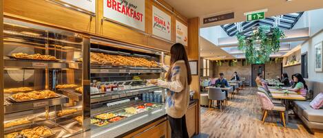 A breakfast buffet with various food options, a seating area with tables and chairs, and a green hanging plant in the center.