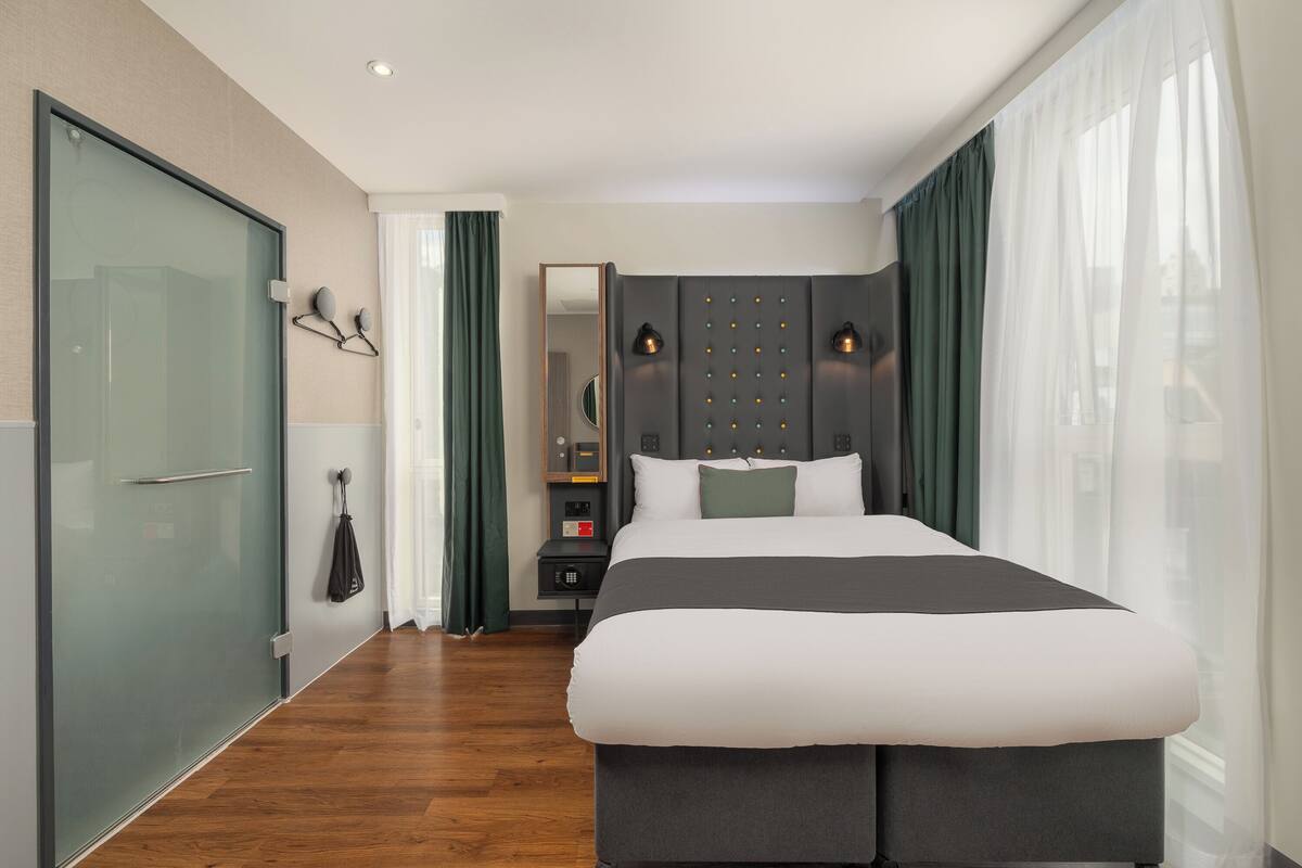 Ein modernes Hotelzimmer mit einem großen Bett, einer Duschkabine aus Glas, einem Schminktisch mit Spiegel und einem Nachttisch.
