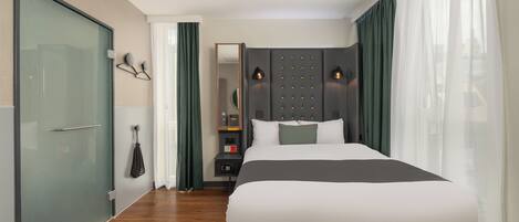 A modern hotel room with a large bed, a glass shower enclosure, a vanity with a mirror, and a nightstand.