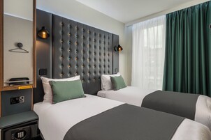 Ein Hotelzimmer mit zwei Betten, einem gepolsterten Kopfteil, Nachttischlampen, einem Spiegel, einem Kleiderständer und Blick auf die Stadt durch große Fenster.
