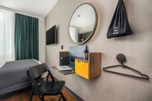 A hotel room with a bed, a desk with a laptop, a mirror, and a hanging bag labeled "Hot Stuff".