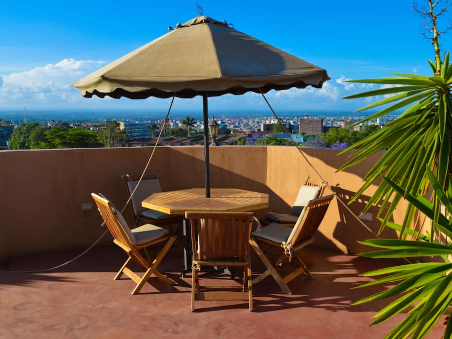 Una terraza con una mesa redonda de madera y cuatro sillas, un paraguas color beige, y vistas a un paisaje urbano.
