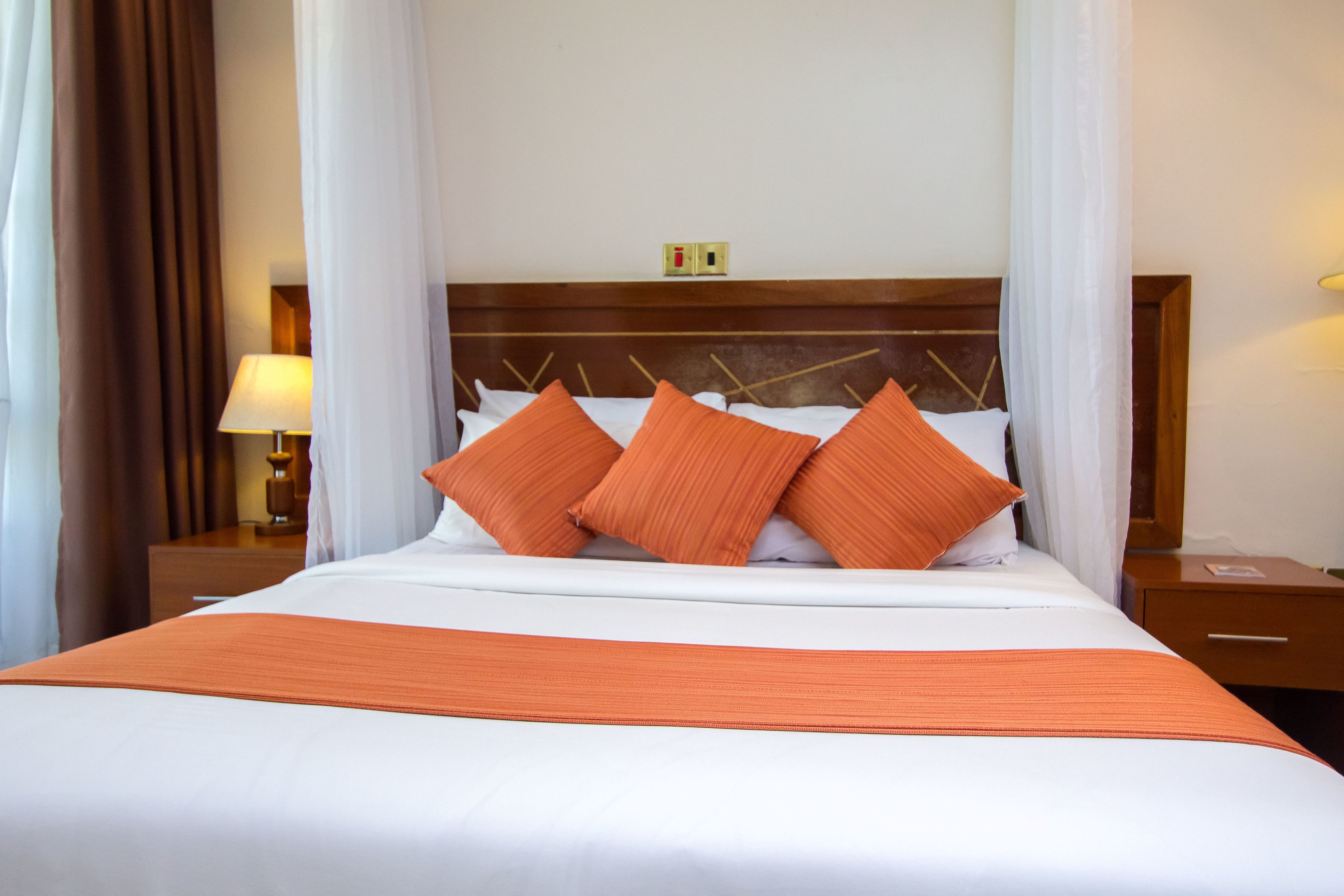A neatly made bed with white bedding and orange pillows, a wooden headboard, and a bedside table with a lamp.