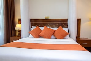 Una cama bien hecha con ropa blanca y almohadas naranjas, un cabecero de madera y una mesita de noche con una lámpara.