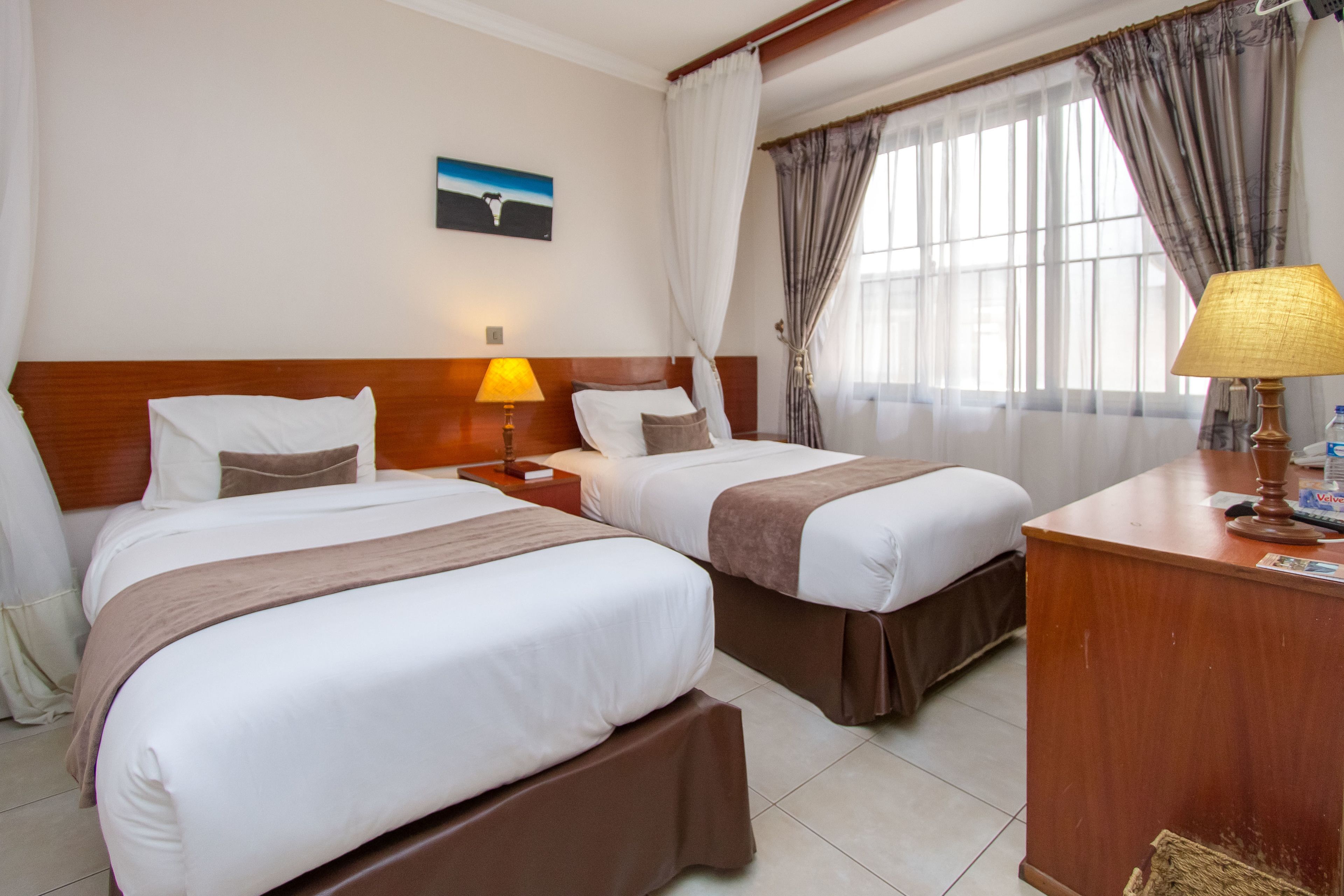 A hotel room with two beds, a desk, a lamp, and a painting on the wall.