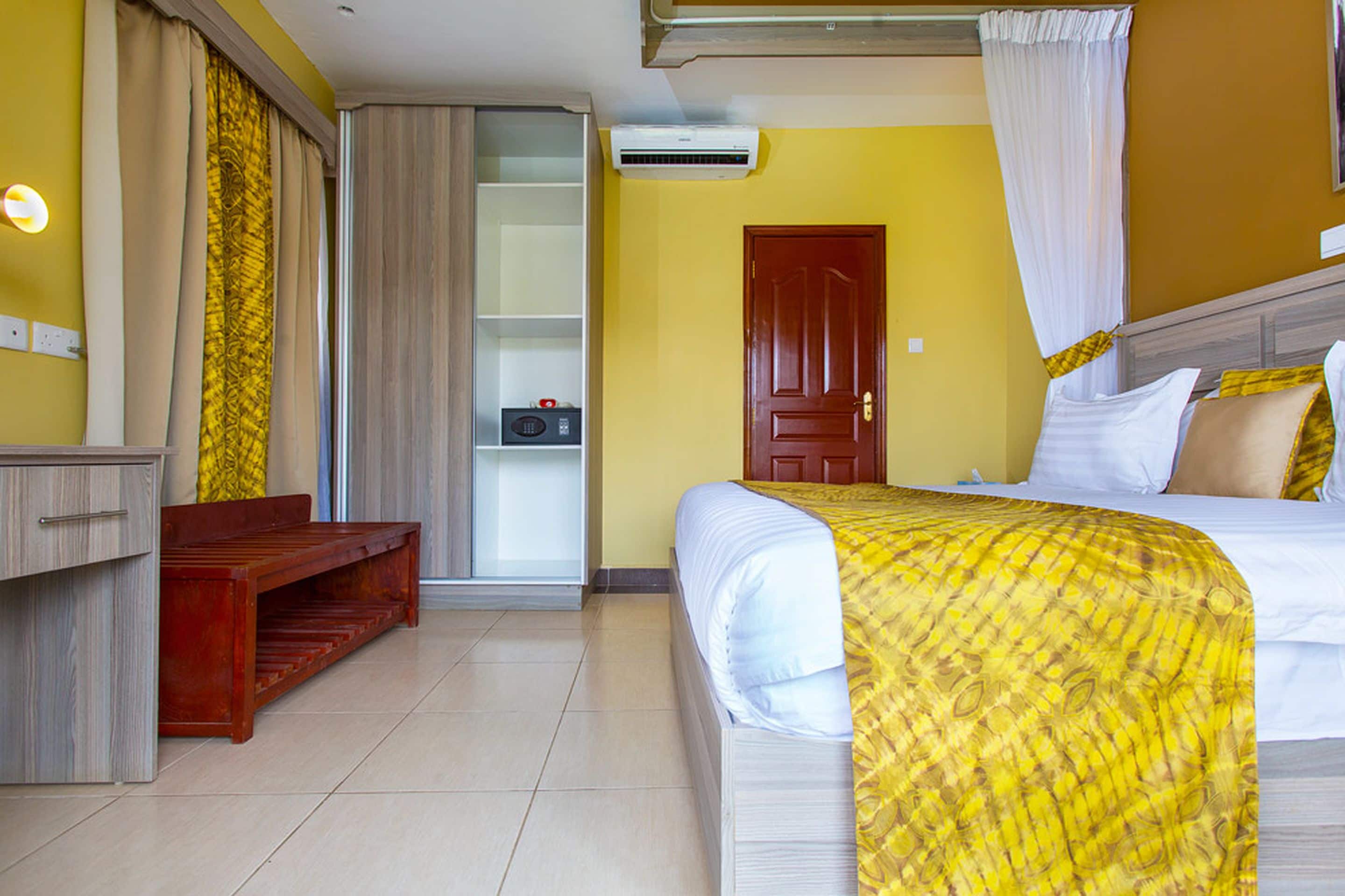 A hotel room with a bed, a wooden bench, a wardrobe, a door, and an air conditioning unit.