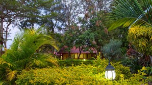 A lush garden with a variety of plants and a traditional building with a red roof in the background. - Wigot Gardens (Kisumu)