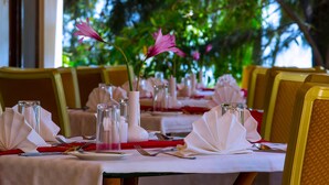 A restaurant setting with tables set for dining, featuring white tablecloths, red napkins, and clear glass vases with pink flowers. - Wigot Gardens (Kisumu)