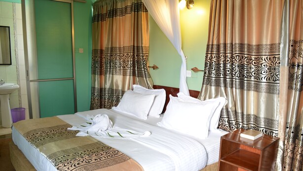 A neatly made bed with white linens and a decorative headboard, a bedside table with a book, and a bathroom with a sink and mirror. - Royal City Hotel (Kisumu)