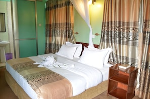 A neatly made bed with white linens and a decorative headboard, a bedside table with a book, and a bathroom with a sink and mirror. - Royal City Hotel (Kisumu)