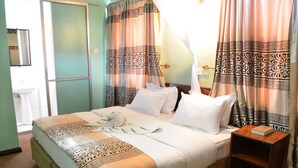 A neatly made bed with white linens and a decorative blanket, a bedside table with a book, and curtains with a patterned design. - Royal City Hotel (Kisumu)